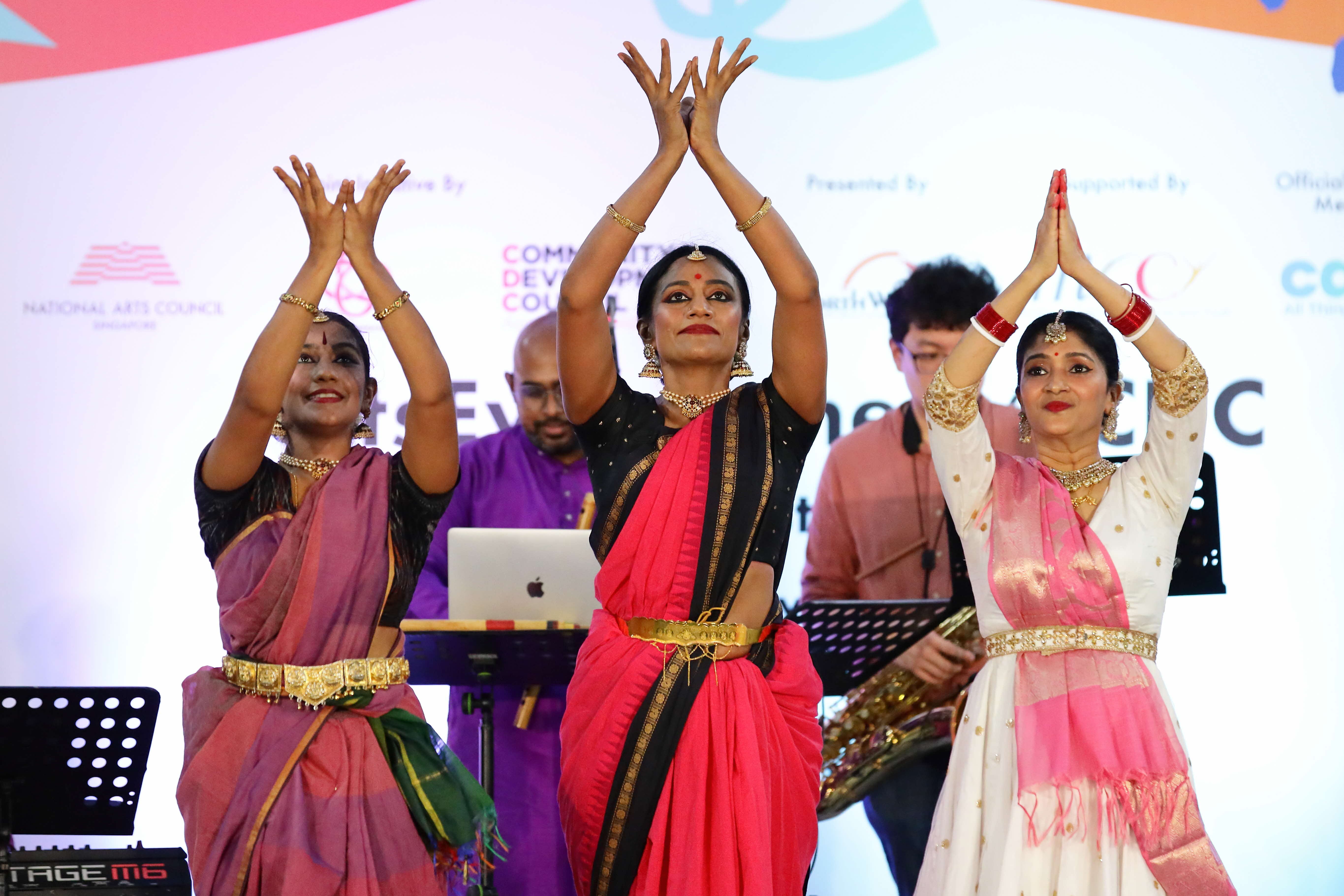 Three women in traditional Indian clothing dance on stage. Behind them are musicians and sponsor logos.
