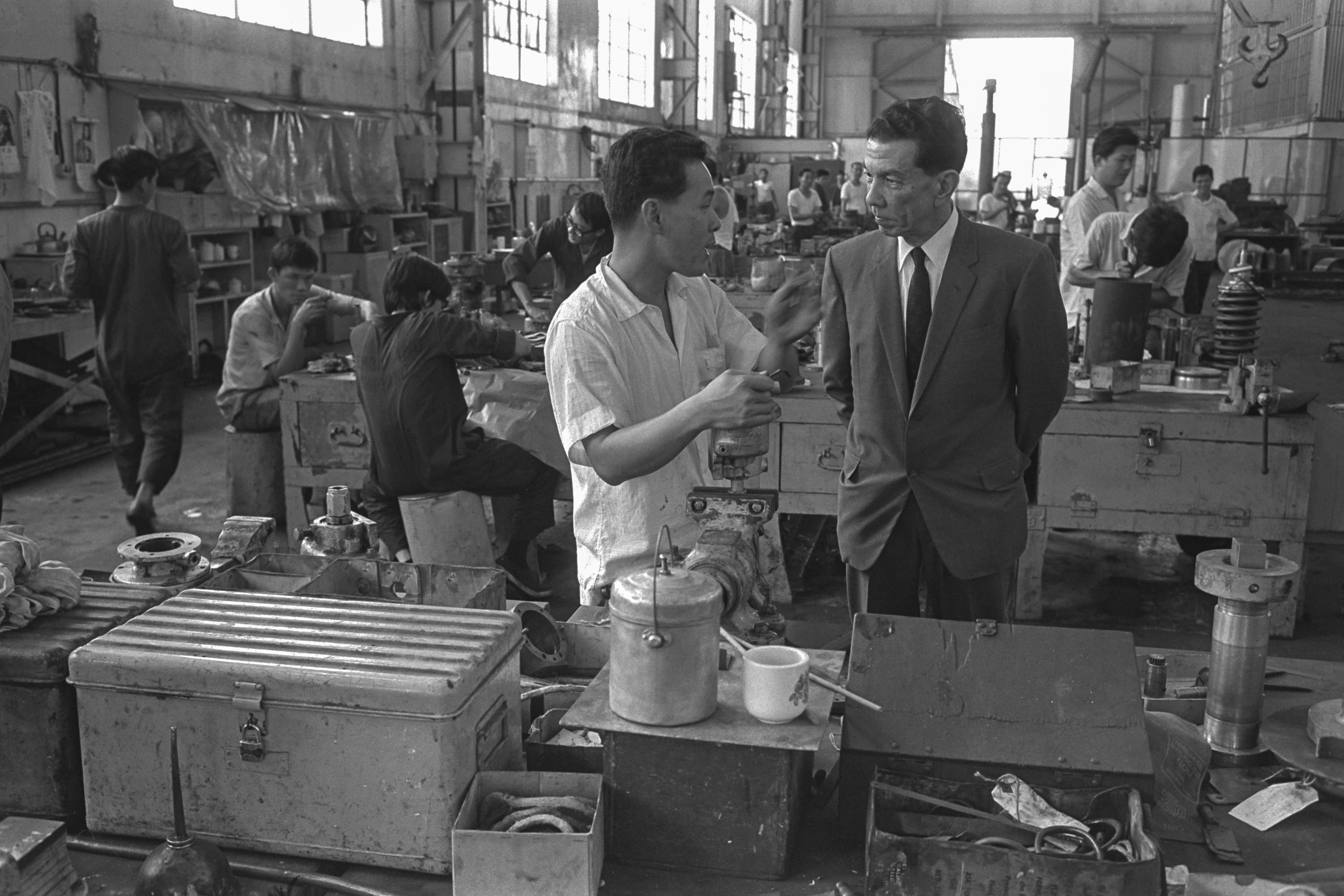 Black and white photo of a factory interior with workers. A man in a suit speaks to a worker in a white shirt.