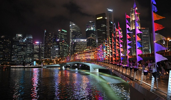 Night view of Singapore skyline with lit bridge decorated with colorful flags reflecting in the water.