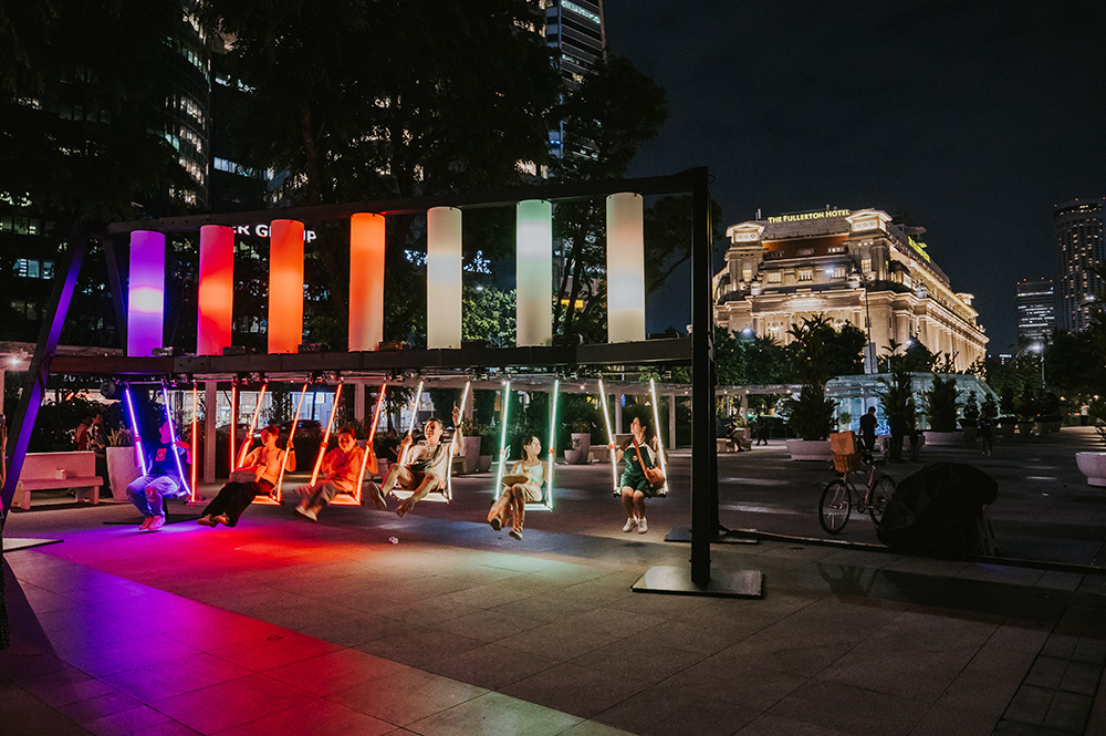 People on LED swings, lit in rainbow colors, in an urban park near The Fullerton Hotel at night.