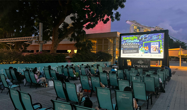 Outdoor cinema in Singapore. People are seated in rows watching a movie on a large screen. Marina Bay Sands is in the background.