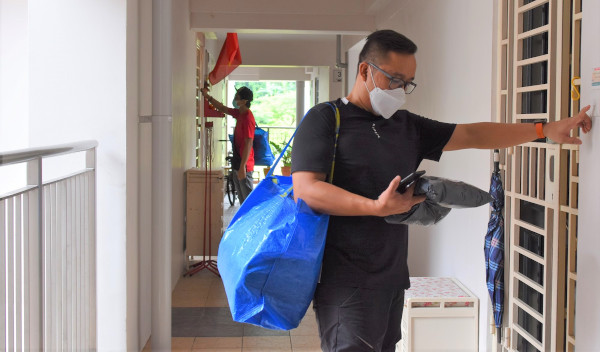 A man with a blue bag rings a doorbell while another hangs red flags in the background. Both wear face masks.