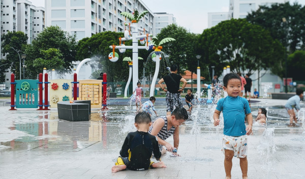 Children play in a splash park with spraying water features and a playground in the background.