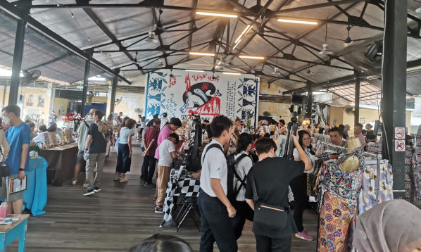 Indoor market with vendors and shoppers browsing stalls with clothes and handmade items. Large graphic banner in the background.