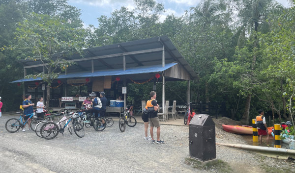 Outdoor activity center. People with bikes, kayaks and a small open-air shop area with red lanterns. Trees and water are visible.