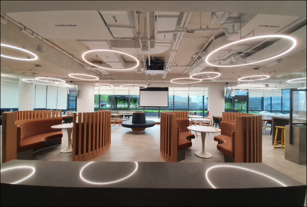 Open office space with booth seating, tables, and a projection screen. Modern, circular light fixtures hang from the ceiling.