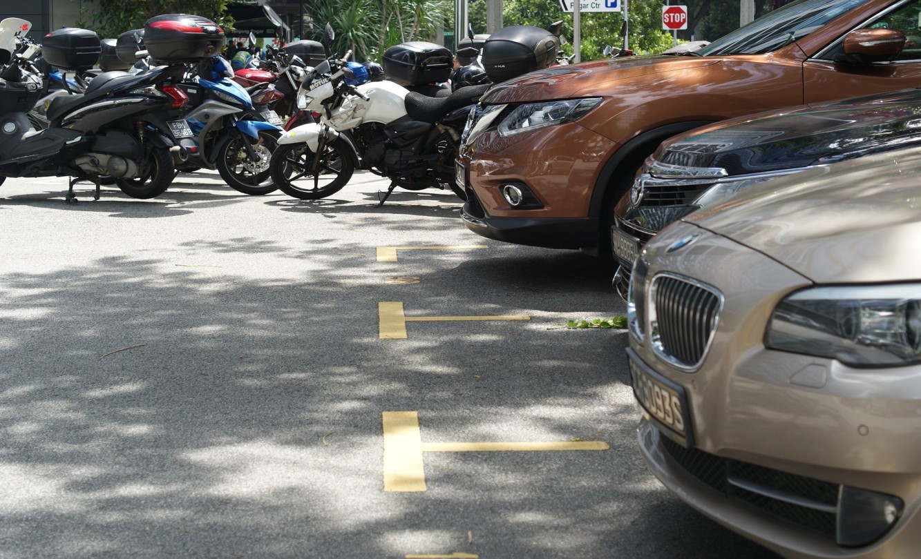 Motorcycles and cars parked in marked spots on paved lot with tree shadows.