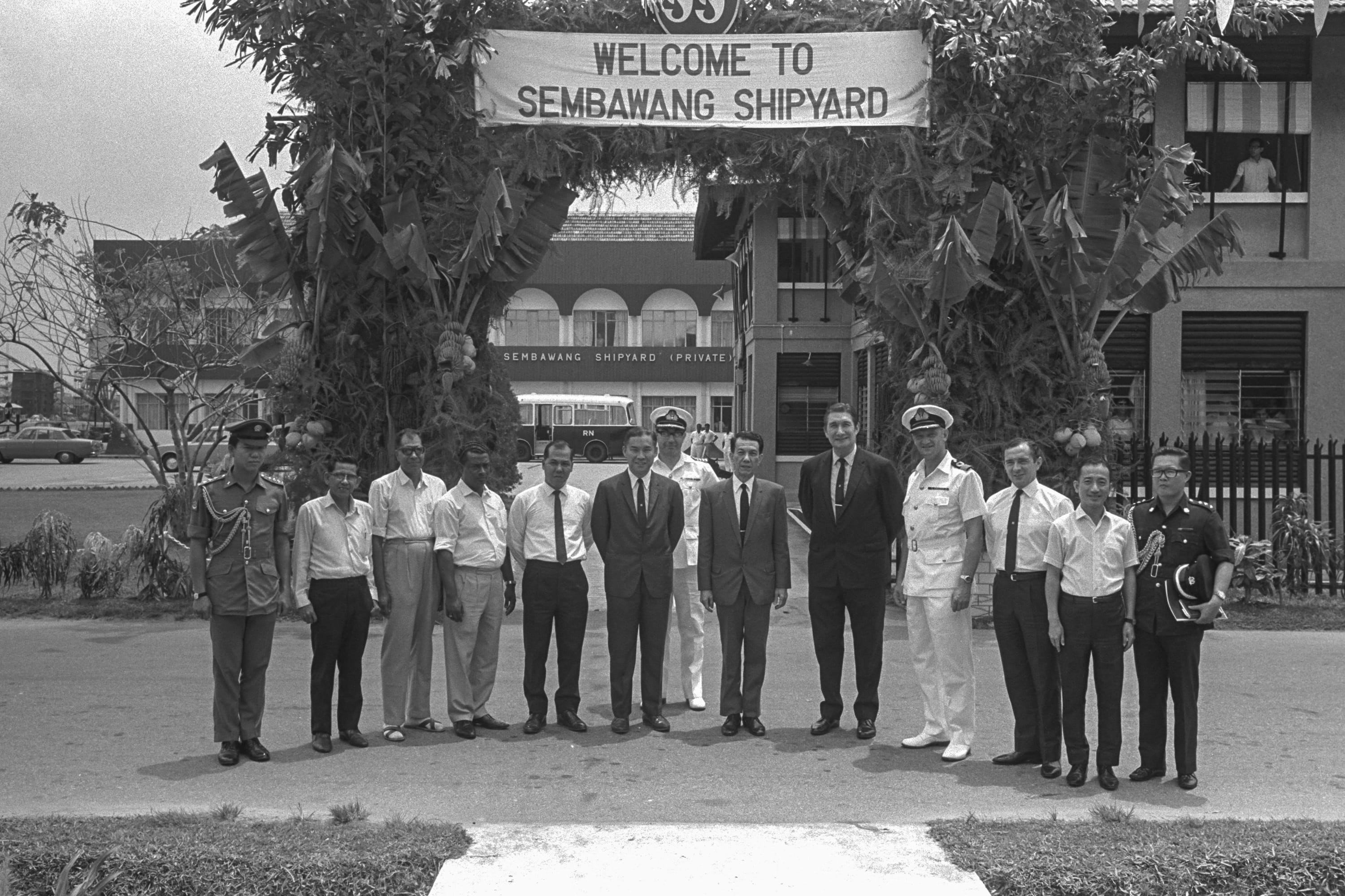 Black and white photo of a group of officials standing under a "Welcome to Sembawang Shipyard" sign.