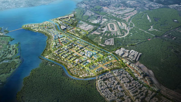 Aerial view of a planned community with buildings, green spaces, and a racetrack, bordering water and forests.