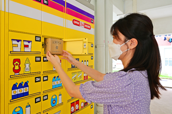 A woman retrieves a box from a yellow parcel locker with illustrations of goods. She wears a mask.