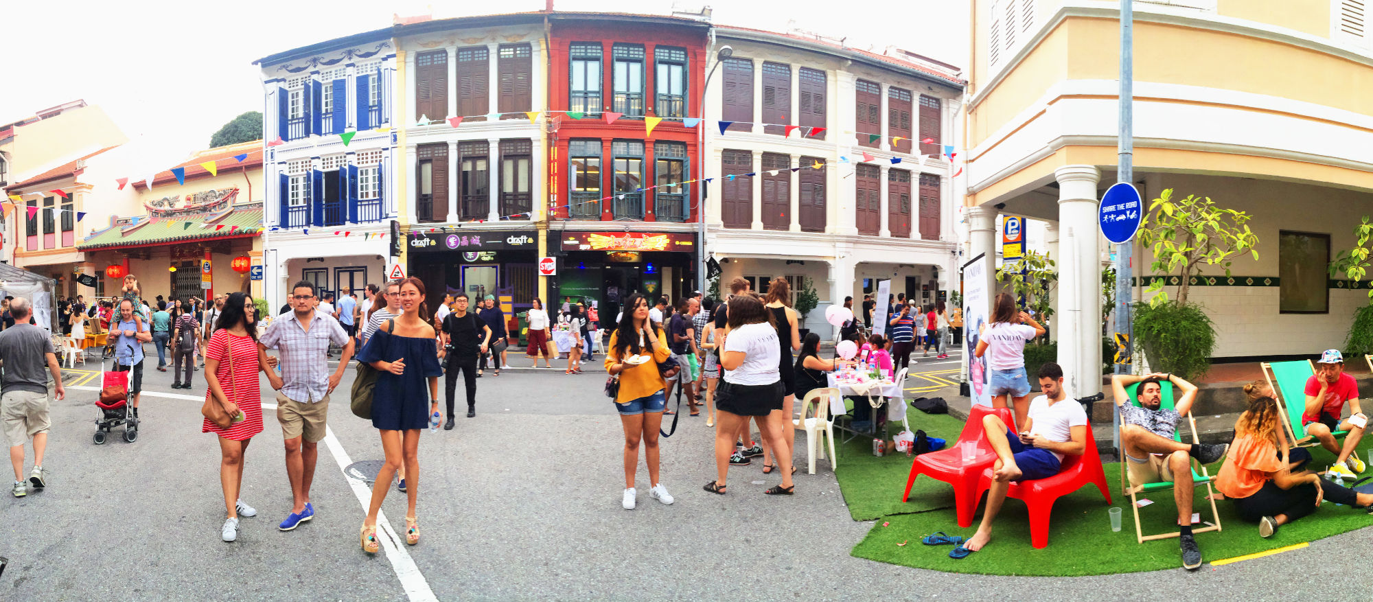Street scene with crowds, shophouses in colors, and seating area on astroturf.