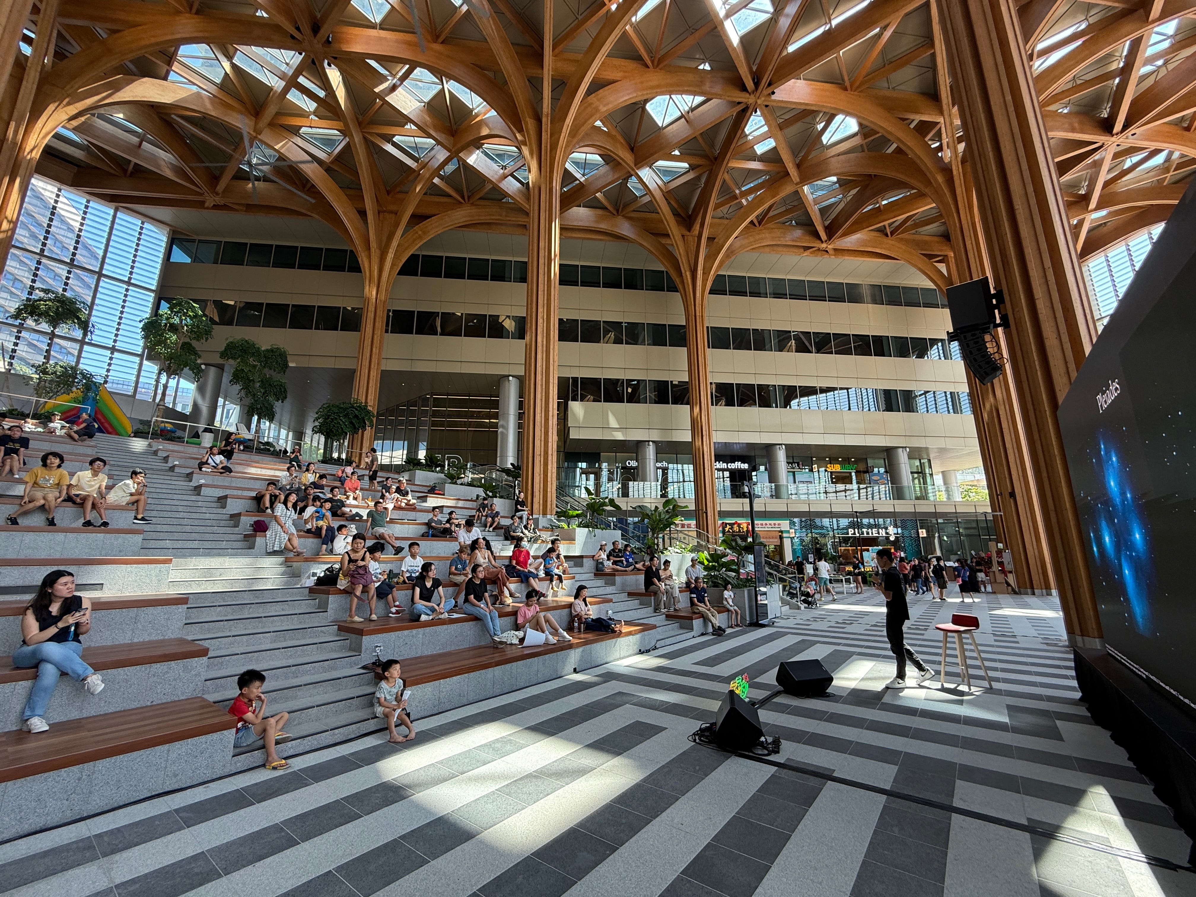 Open-air auditorium with wood canopy and bleacher seating filled with people. Speaker on stage.