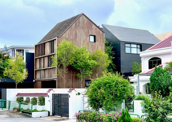 A modern wooden house with a unique architectural design stands beside other contemporary houses, surrounded by greenery and neatly trimmed hedges.