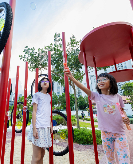 Two girls with glasses playing with bubbles at a playground with red posts and bicycle wheels.