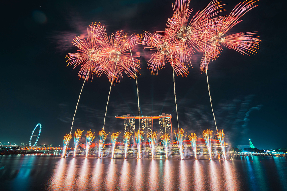 Fireworks over water at night with Marina Bay Sands and Ferris wheel in the background.