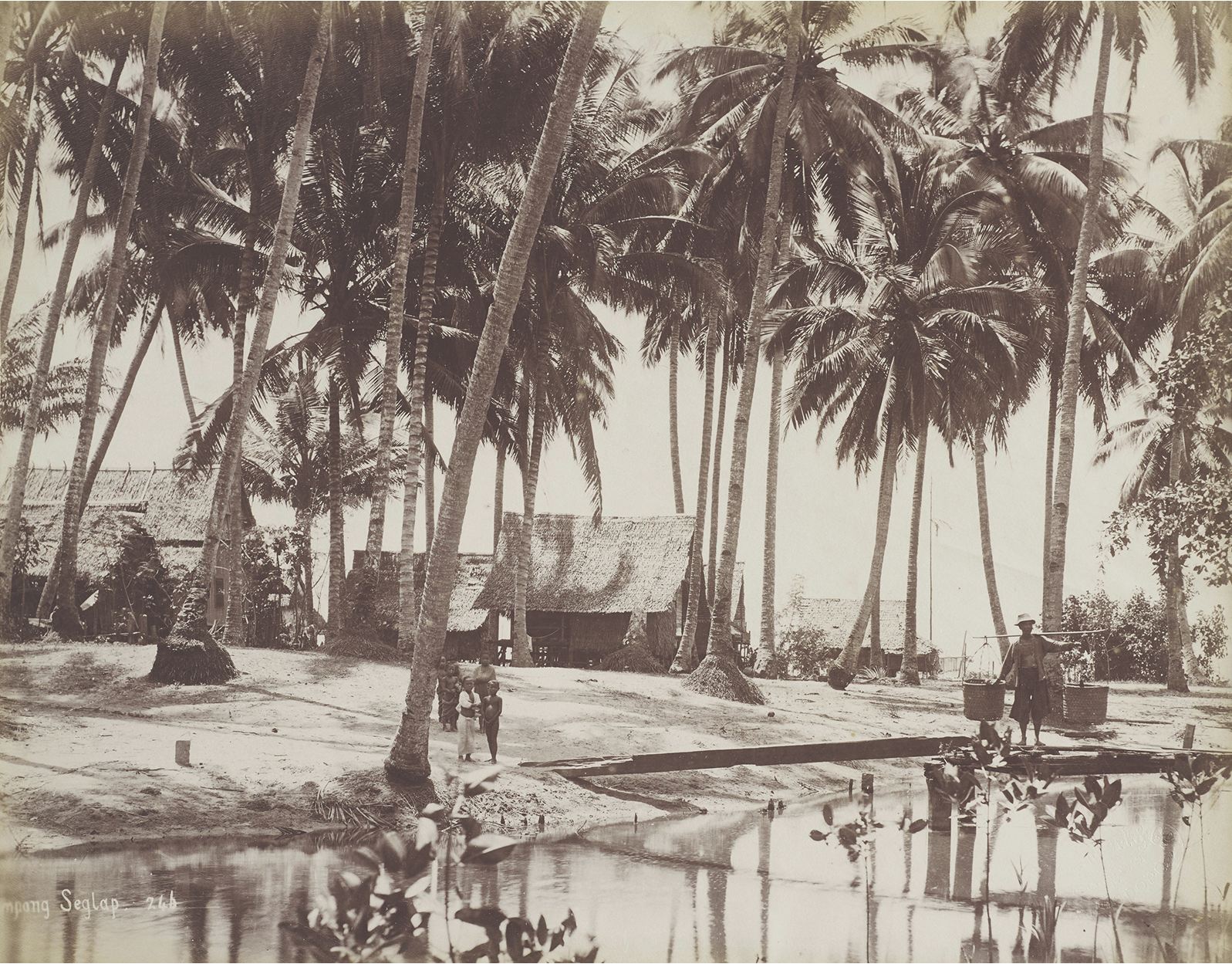 Sepia photo of a tropical village with thatched huts, palm trees, and people crossing a small canal on a narrow bridge.