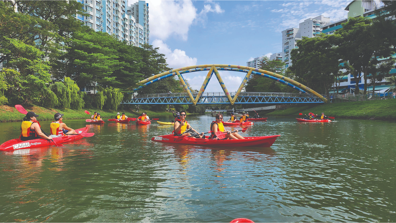 A group of people kayaking on a river under a bridge, with buildings and trees in the background.