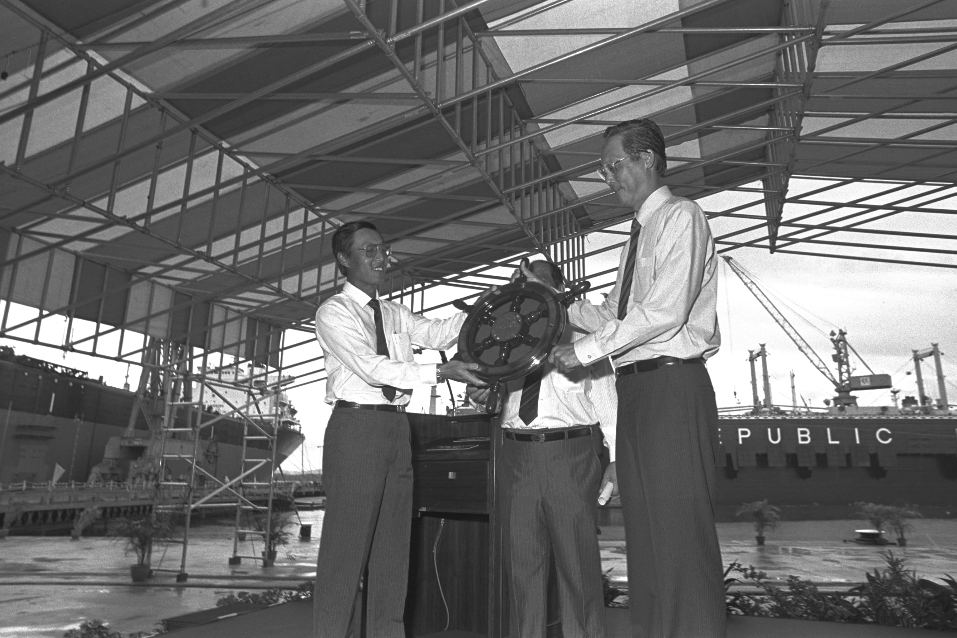 Black and white photo of three men at a shipyard passing a ship's wheel under an industrial structure. "PUBLIC" is visible on a large ship in the background.