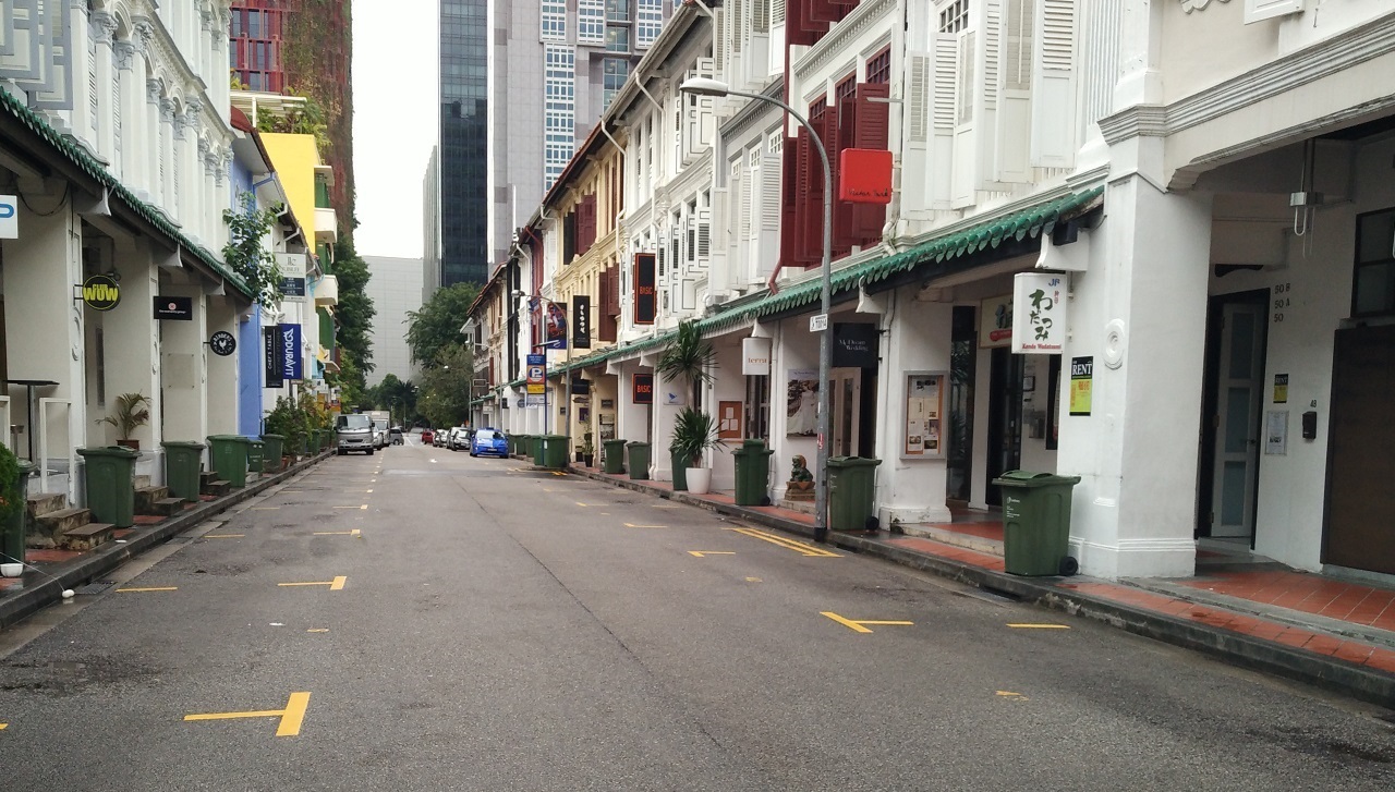 Street view with rows of buildings, sidewalks, parked cars, and yellow markers on asphalt.