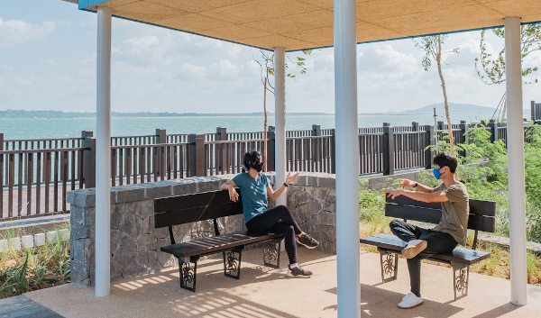 Two masked people sit on benches, distanced, under a shaded structure near a waterfront.