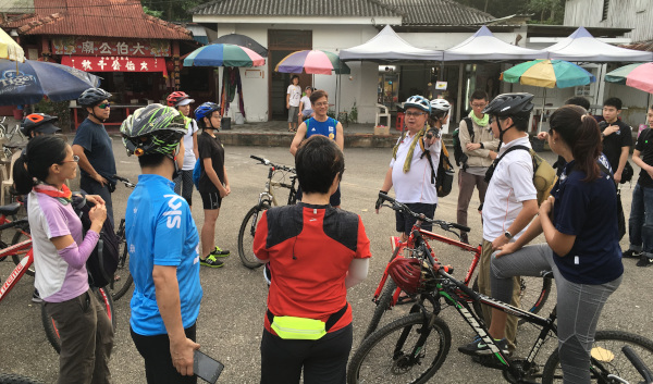 A group of cyclists wearing helmets stand with their bikes, listening to someone speaking. They are in an outdoor area.