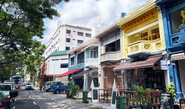 Street view in Singapore with colorful shophouses and traffic. Trees line the sidewalk.