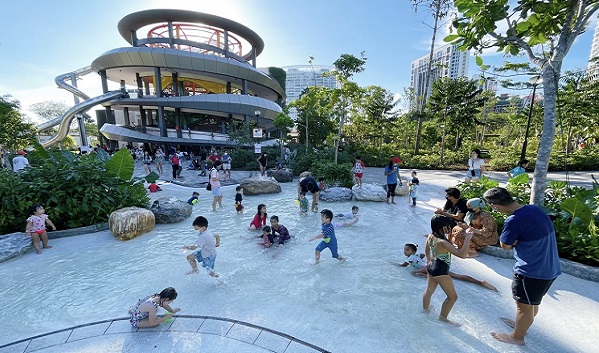 Children play in a shallow water feature at a park with a spiral slide and tower.