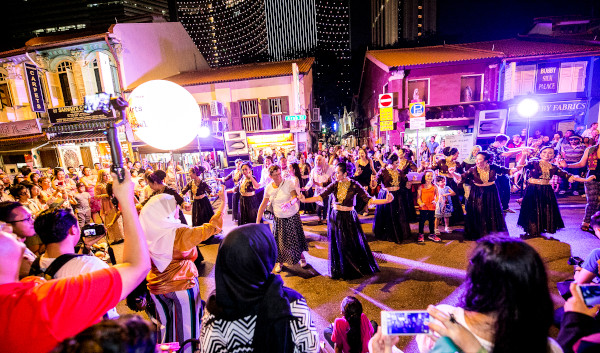 Night street scene with performers in traditional dress dancing for a crowd. City buildings in the background.