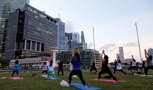 Yoga class on a green lawn with city buildings in the background, all with arms raised.