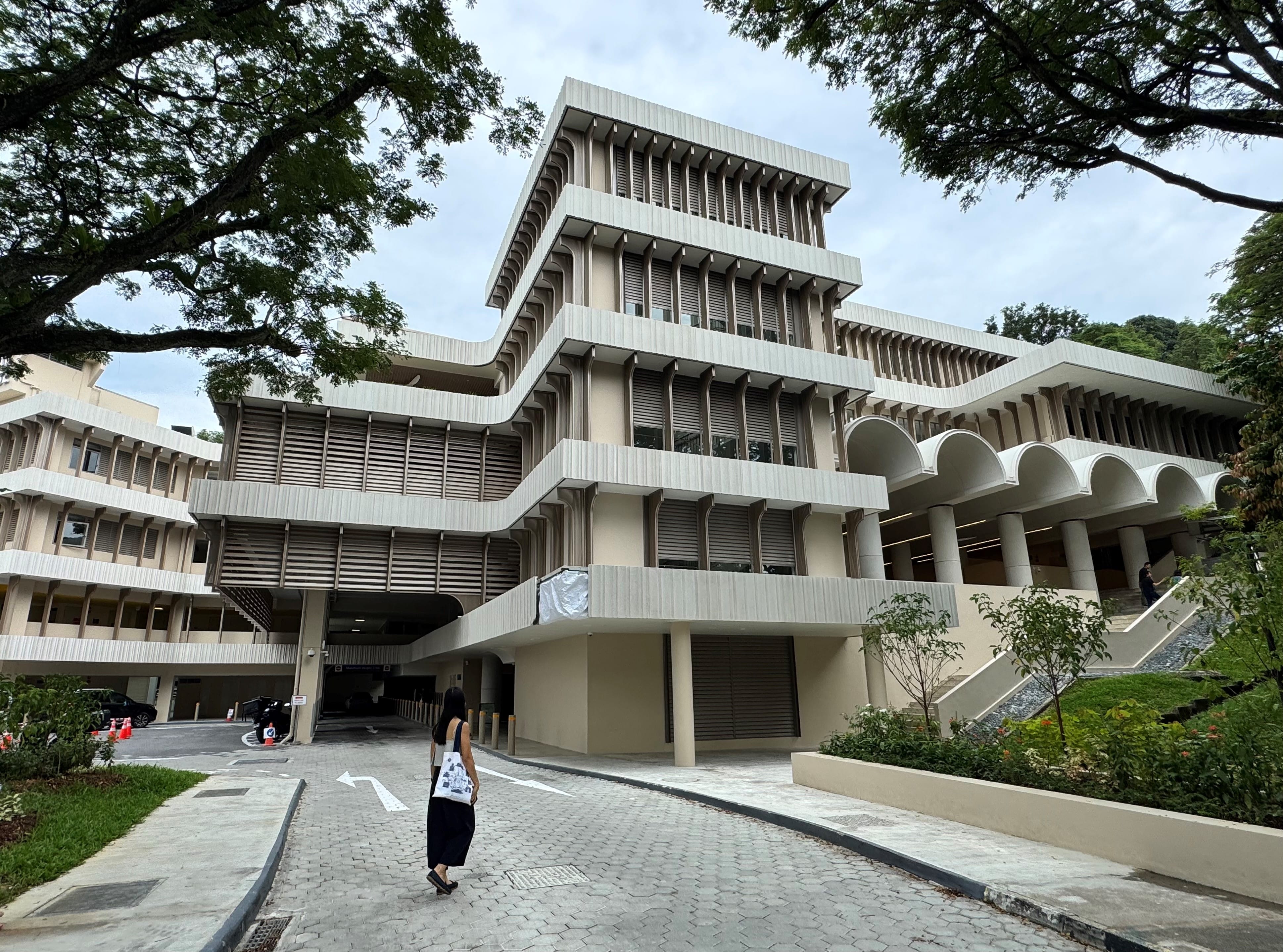 Woman walking towards a multi-story building with horizontal louvers and arched walkways.