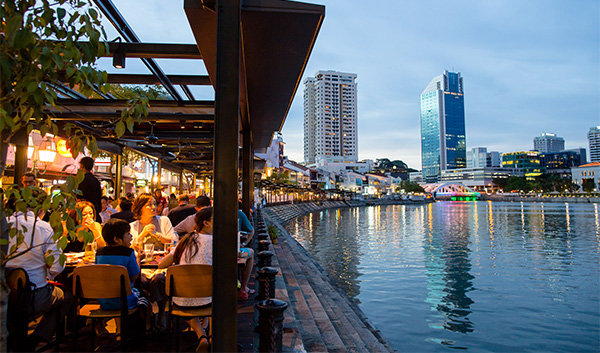 Waterfront restaurant at dusk with city buildings reflecting in river. People dining under awning.