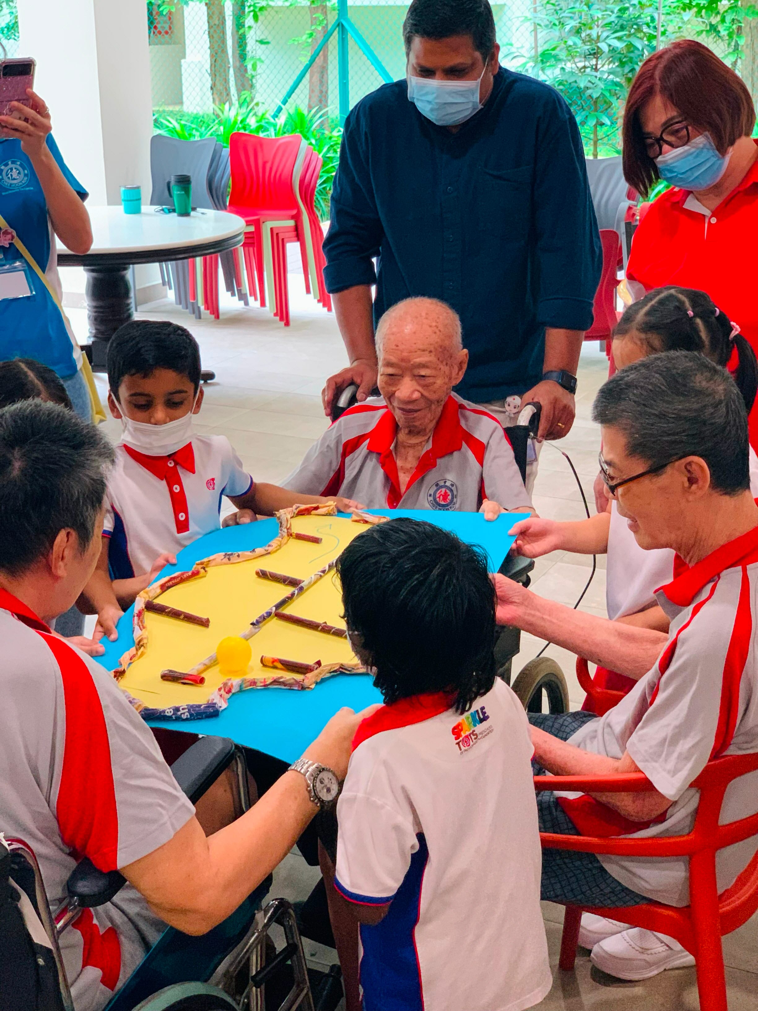 People of various ages gather around a game on a blue board with a yellow surface, some wearing masks. The elderly person sitting in a wheelchair is looking at the game.