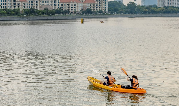 Two people kayak in an orange boat on a river. Buildings are in the background.