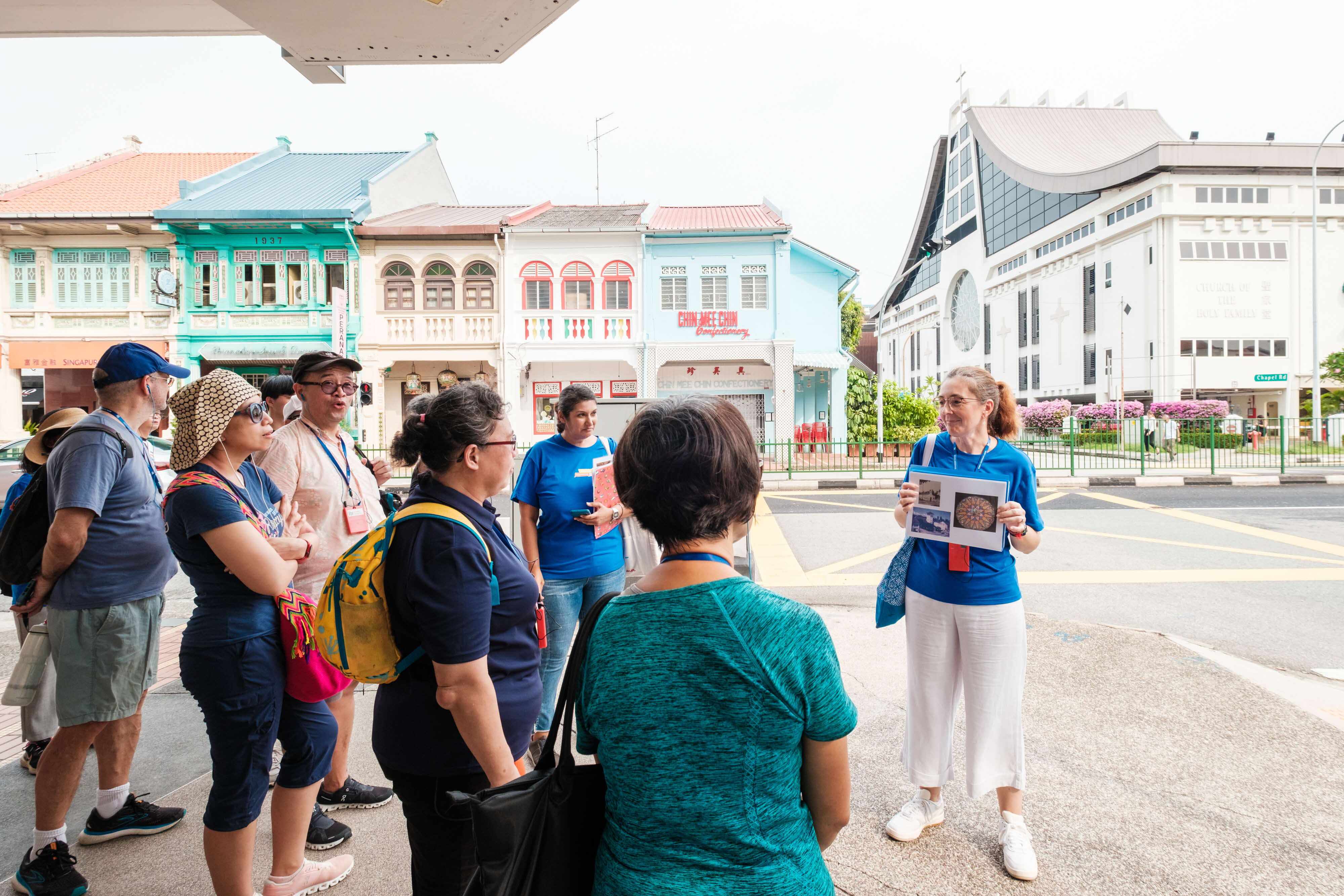 A tour guide presents photos to a group in front of colorful buildings and a church in Singapore.