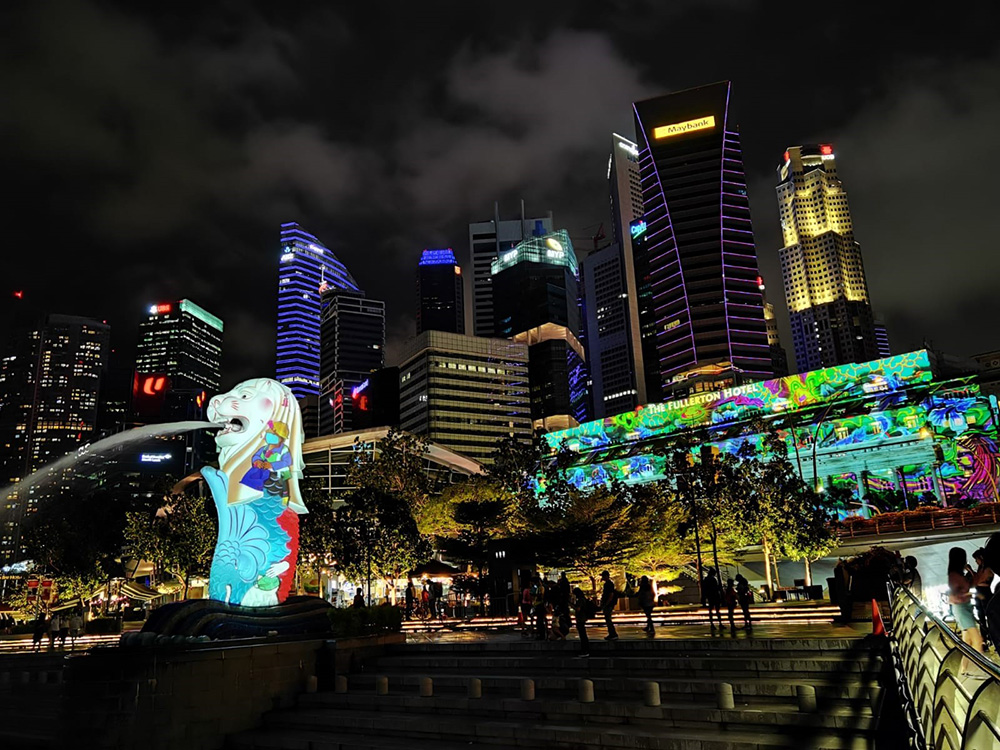 Night view of Singapore skyline; Merlion statue with water jet, colorful building illuminations.