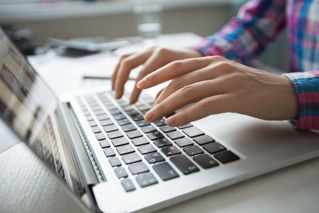 Hands typing on a silver laptop keyboard, wearing a blue and pink plaid shirt.