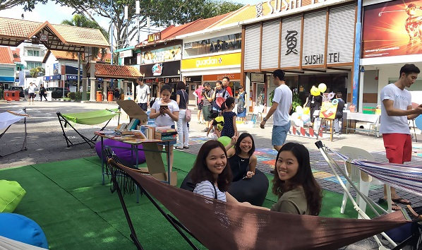 Outdoor community event with people, shops, hammocks, and beanbag chairs. "Sushi Tei" restaurant in the background.