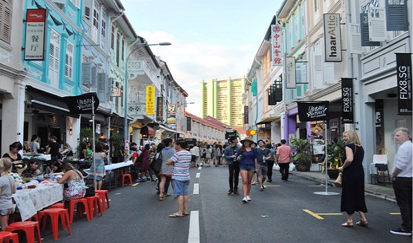 Street scene in Singapore with people walking and eating at outdoor tables along colorful buildings.