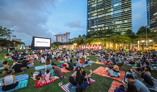 Outdoor movie night with a large crowd on blankets watching "Carnival by the Bay" on screen.
