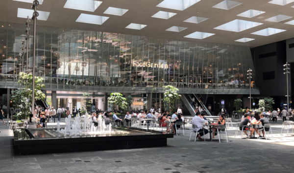 Indoor courtyard with people dining, a fountain, plants, and "Food Garden" sign on a glass building.