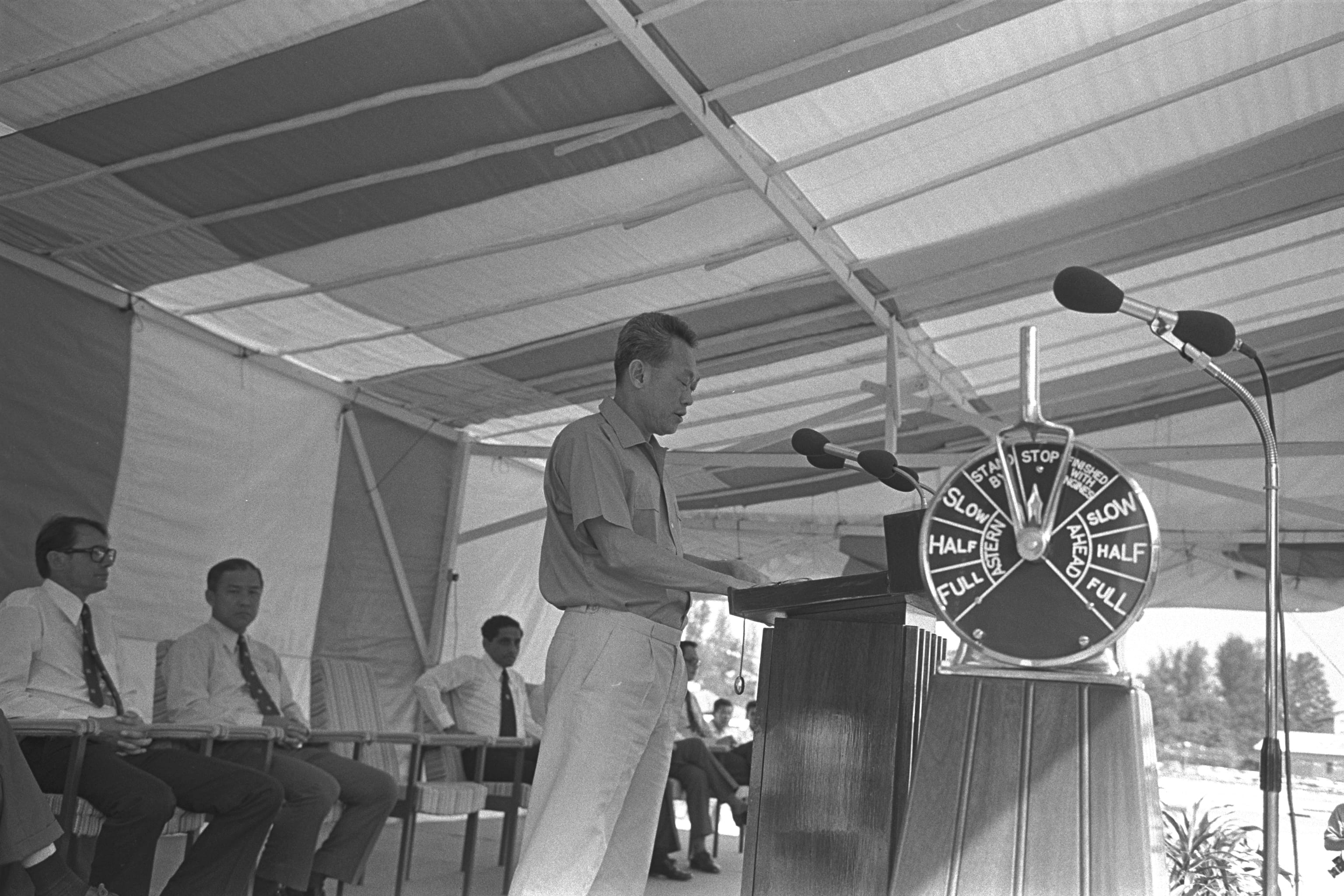 Black and white photo of Lee Kuan Yew speaking at a podium in 1969. He is surrounded by other people.
