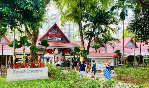 Newton Food Centre in Singapore, surrounded by greenery and people. The hawker centre has a distinctive red-roofed structure.