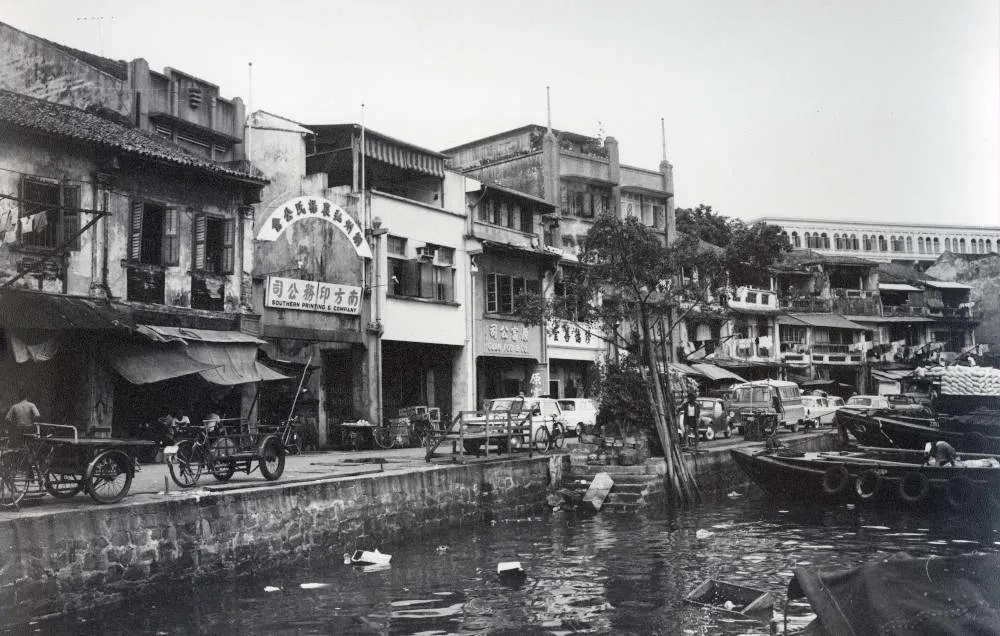 Black and white photo of a waterfront street lined with buildings, carts, cars, and boats.