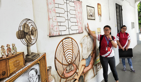 Two women stand in front of a wall mural depicting a vintage home interior with furniture and decor.