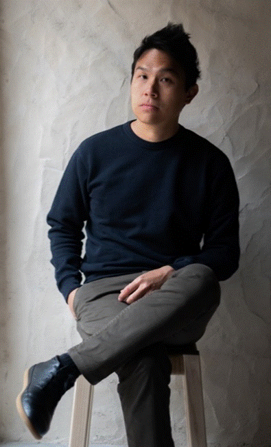 Portrait of a man seated on a stool against a textured grey wall, wearing a dark navy sweater and grey trousers.