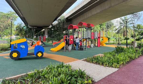 A colorful playground under a highway overpass features slides, climbing structures, and a blue train play structure. People are playing and standing near the equipment.