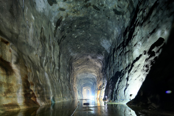 A flooded tunnel with rough-hewn rock walls leads to a distant light.