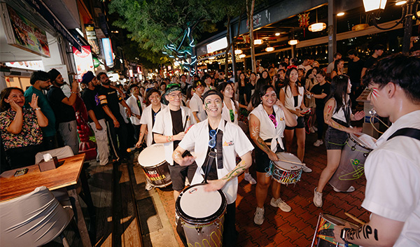 Street scene at night. People with face/arm paint are playing drums, and a crowd is gathered around them.