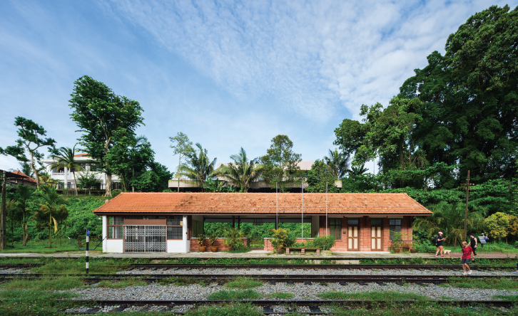 “Small rustic railway station with orange tiled roof beside train tracks, surrounded by lush greenery under a bright blue sky.
