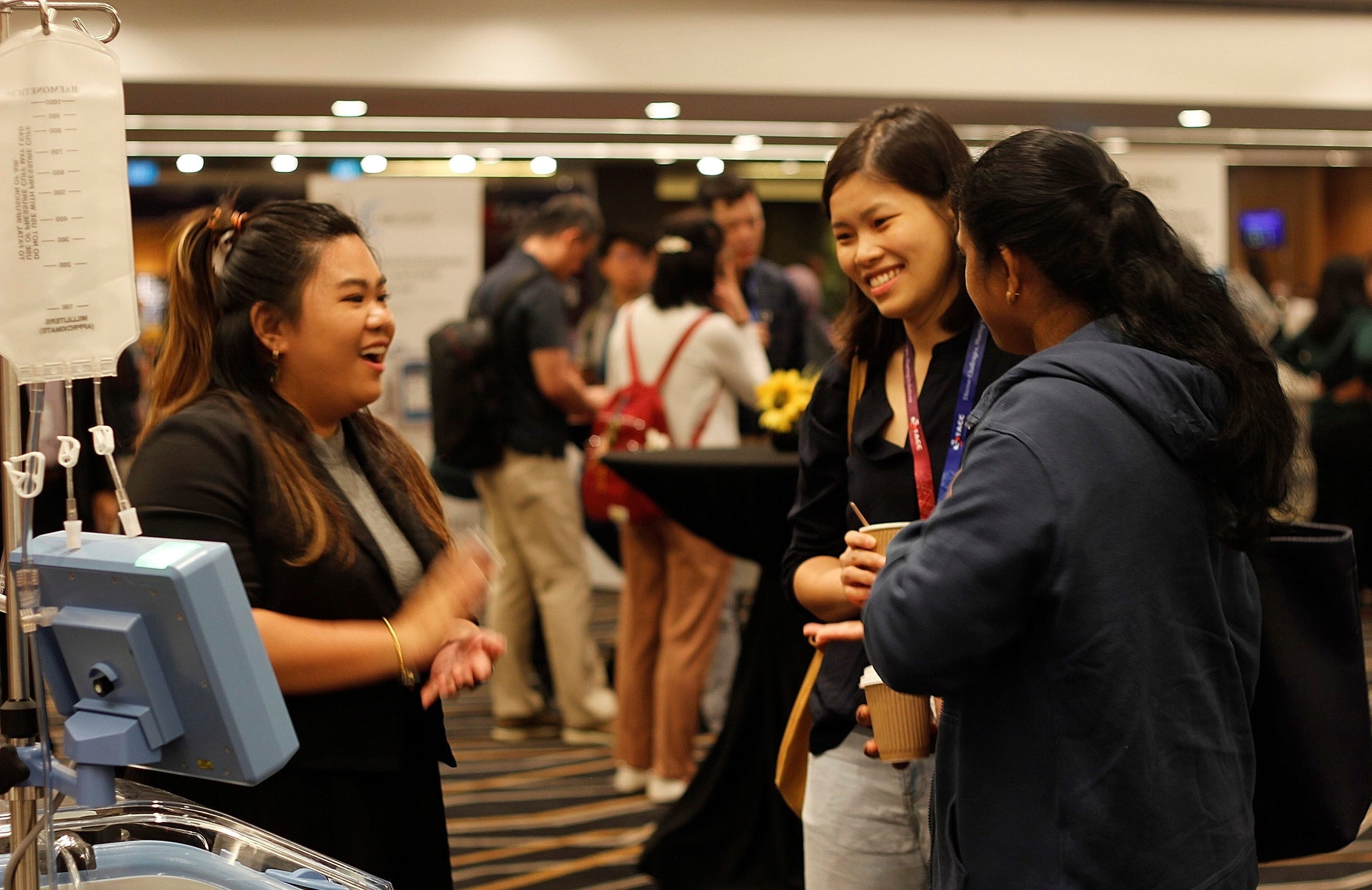 Three women smile and chat at an indoor exhibition booth with medical equipment nearby and attendees in the background.